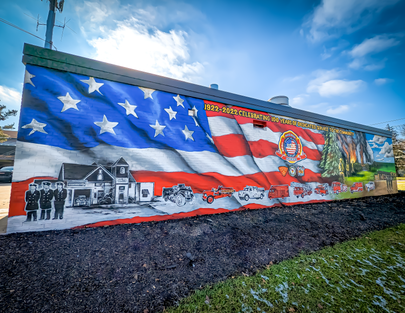 Landscape exterior photograph of a large mural painted on the side of a building housing the South Euclid Fire Department. The mural celebrates the department's 100 year anniversary and depicts fire trucks, fire fighters, and logos on a large American flag background.